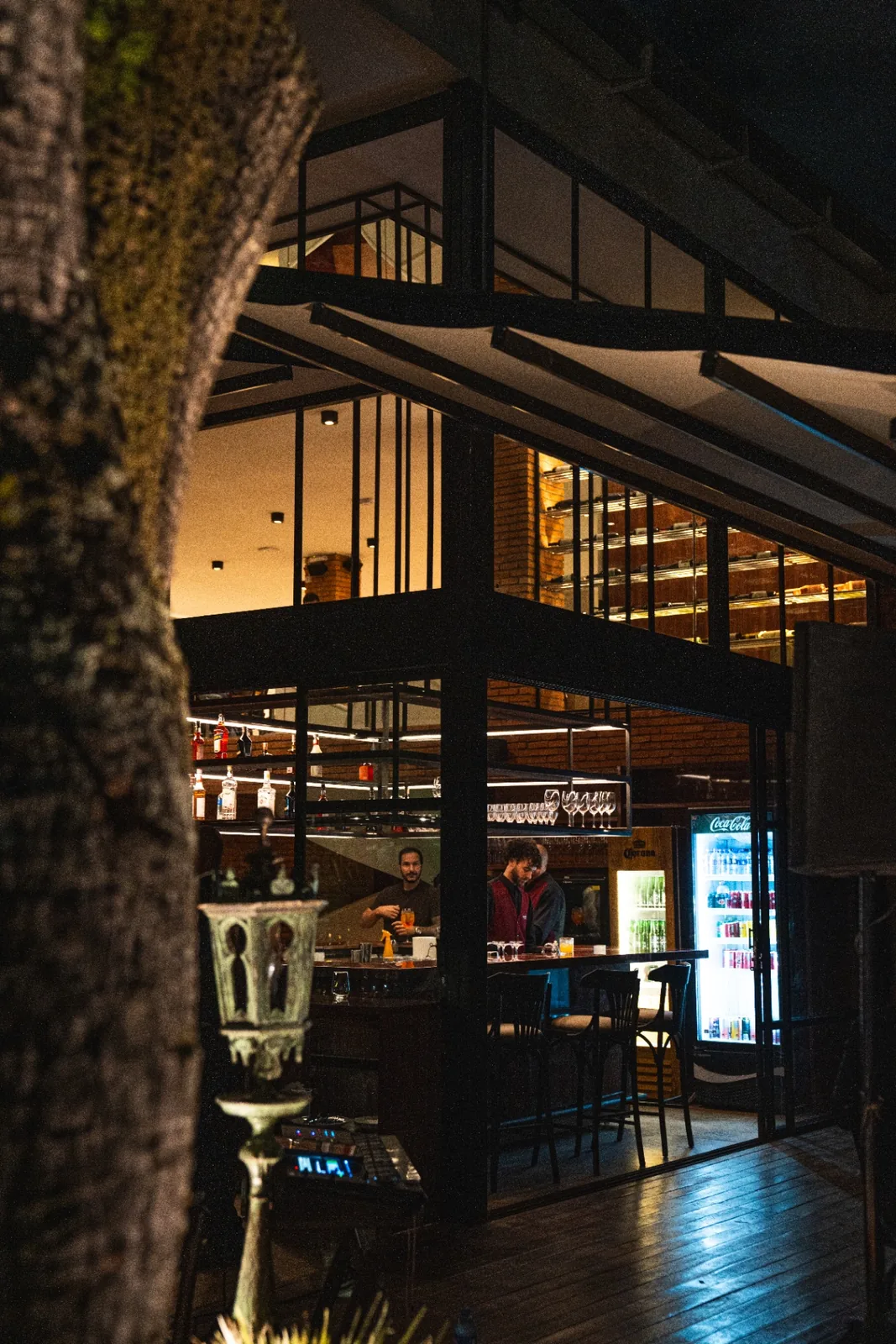 Casa da Albero à noite, vista entre árvores — estrutura metálica, adega iluminada e bar em operação.