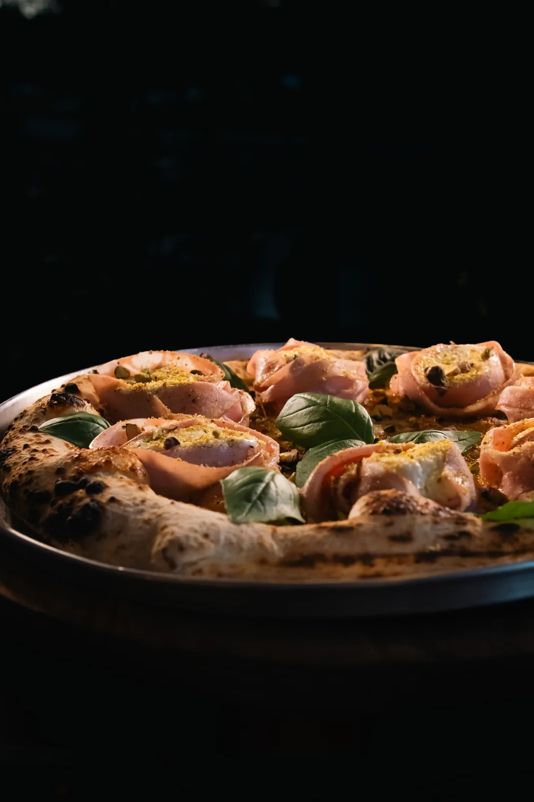 Pizza de mortadella com burrata, pistache e manjericão sobre bandeja preta, fotografada em contraluz.