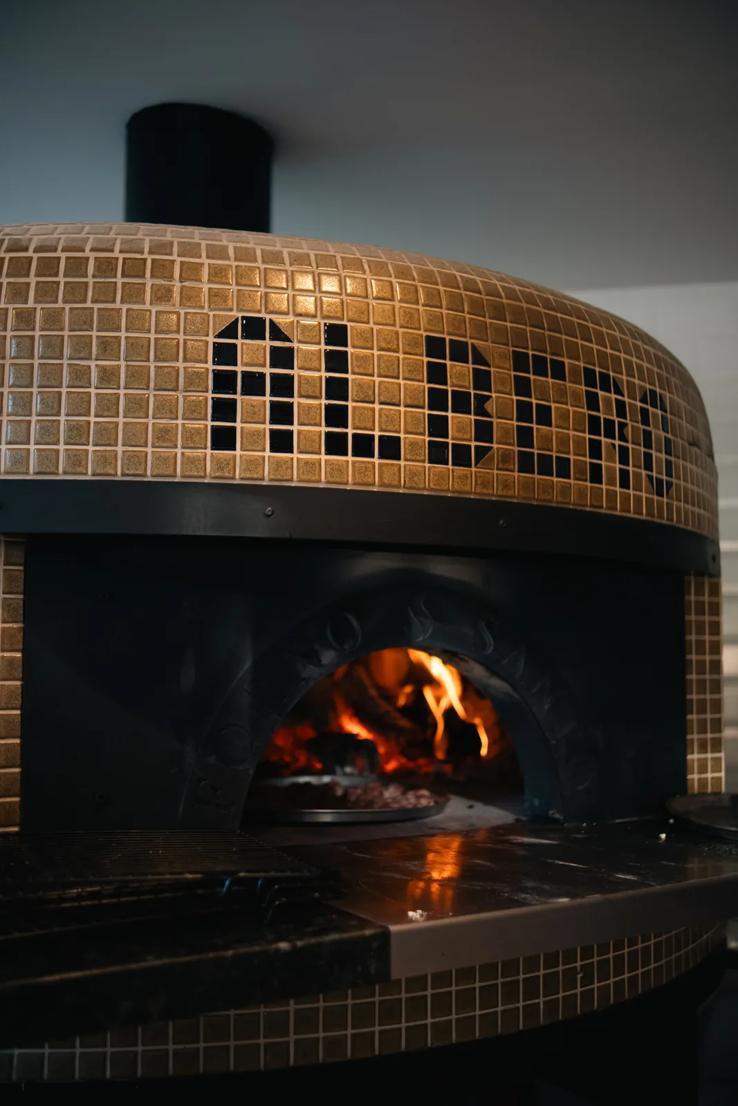 Forno à lenha revestido em mosaico dourado com o nome ALBERO grafado em pastilhas escuras.